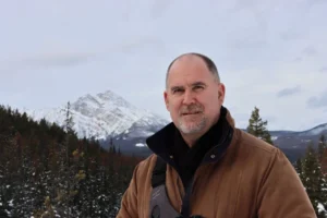 Photographer Bruce Graham casually stands with snow covered mountains in the distant background.